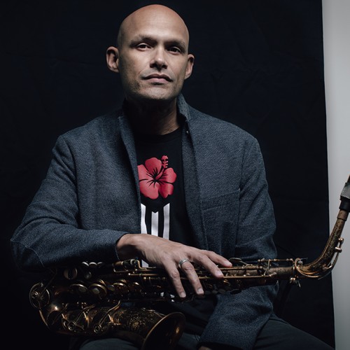 A Puerto Rican man in a gray jacket sits with a saxophone across his lap in front of a black background. 