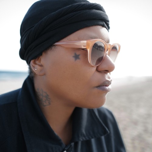 A black woman with a black knit cap, black collared shirt, and sunglasses looks into the distance outdoors. 