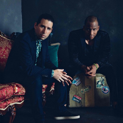 Two Cuban musicians sit in a dark room staring straight ahead, one is on a ornate red couch, one is on a vintage travel suitcase 