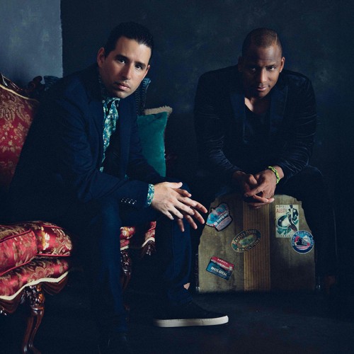 Two Cuban musicians sit in a dark room staring straight ahead, one is on a ornate red couch, one is on a vintage travel suitcase 
