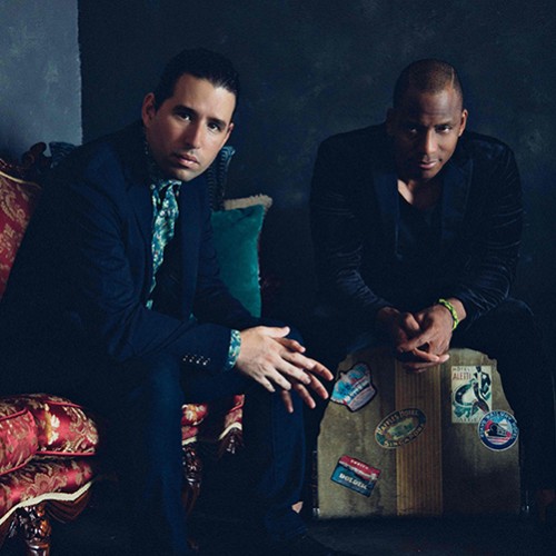Two Cuban musicians sit in a dark room staring straight ahead, one is on a ornate red couch, one is on a vintage travel suitcase 