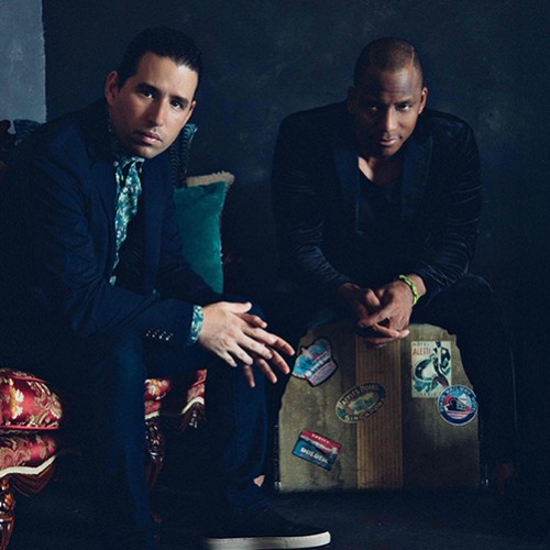 Two Cuban musicians sit in a dark room staring straight ahead, one is on a ornate red couch, one is on a vintage travel suitcase 