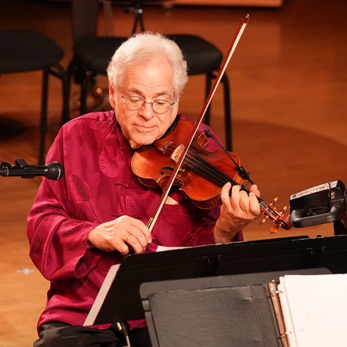 Violinist Itzhak Perlman performing, wearing a deep red shirt and seated on a mobility scooter