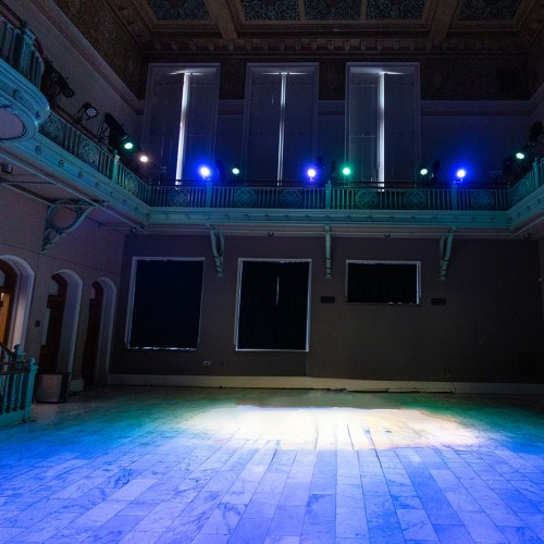 the flat floor of a two-story performance venue in a historic building. The floor is lit with blue, with a white spotlight in the center