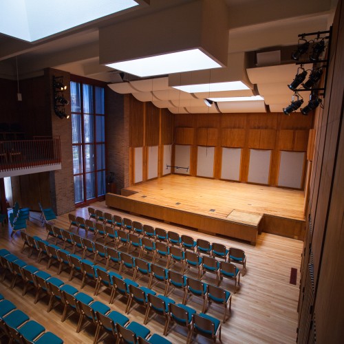 A small, wood paneled auditorium with many blue chairs