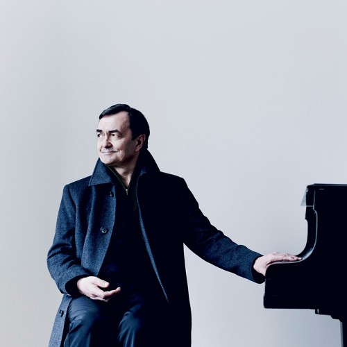 A French man in a long dark coat rests his hands on the piano key lid while looking to the side with a slight smile. 
