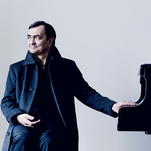 A French man in a long dark coat rests his hands on the piano key lid while looking to the side with a slight smile. 