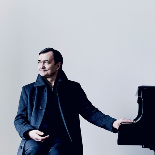 A French man in a long dark coat rests his hands on the piano key lid while looking to the side with a slight smile. 