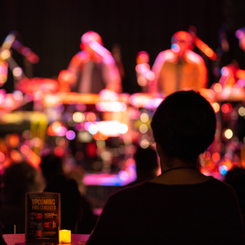 a silhouetted audience at cabaret tables watches a percussion performance on a warmly lit stage