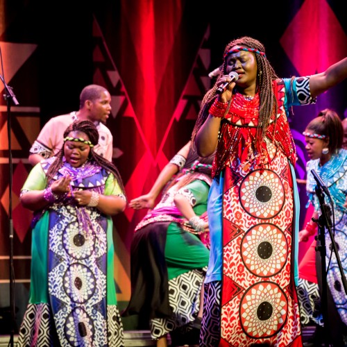 the singers of soweto gospel choir, each wearing a different vibrant color, stand powerfully with fists raised in front of a brownstone building