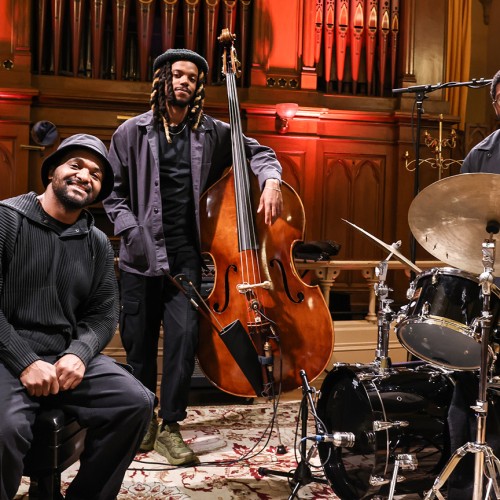 Three black men in black attire stand in front of organ pipes, one sits on a piano bench, the others pose with a bass and drum set. 