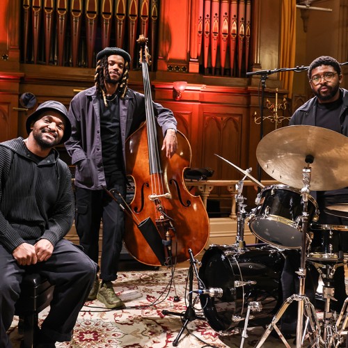 Three black men in black attire stand in front of organ pipes, one sits on a piano bench, the others pose with a bass and drum set. 