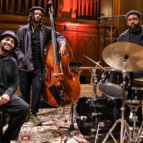 Three black men in black attire stand in front of organ pipes, one sits on a piano bench, the others pose with a bass and drum set. 