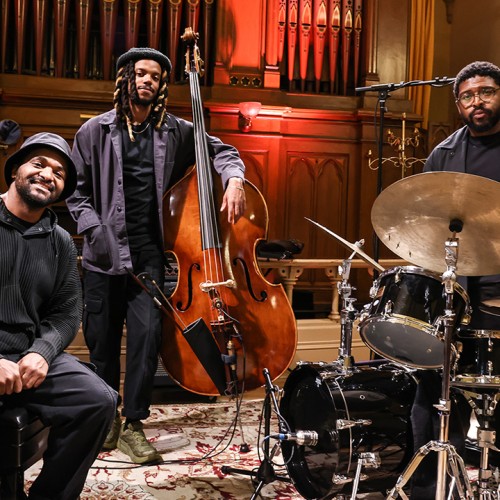 Three black men in black attire stand in front of organ pipes, one sits on a piano bench, the others pose with a bass and drum set. 