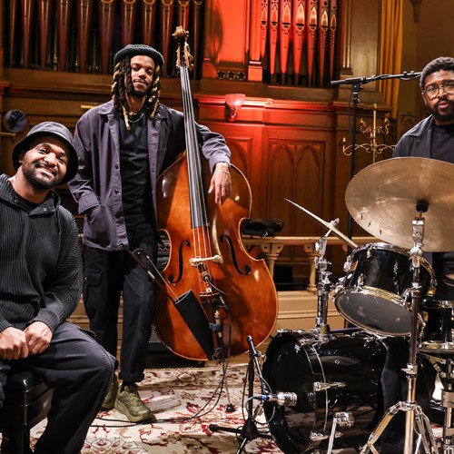 Three black men in black attire stand in front of organ pipes, one sits on a piano bench, the others pose with a bass and drum set. 