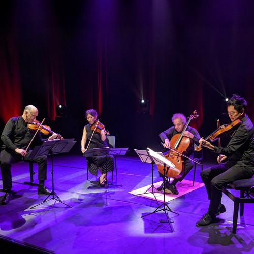 A string quartet of three men and one woman performs on a modern stage with uplighting and purple hues. 