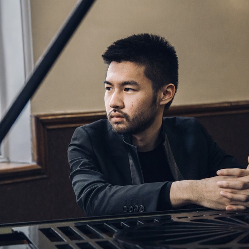 An Asian American man in a black jacket sits leaning on a grand piano, his face framed by the lid and the stick in a well-lit room. 