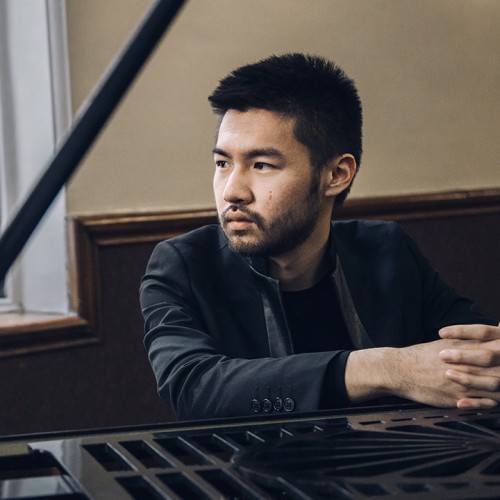 An Asian American man in a black jacket sits leaning on a grand piano, his face framed by the lid and the stick in a well-lit room. 