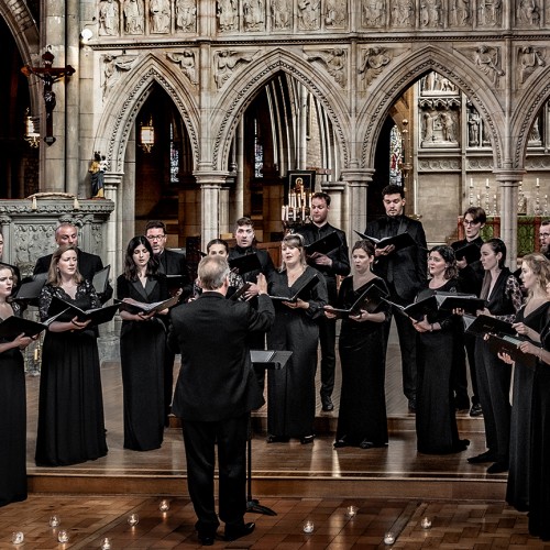 A choir of twenty-one members stand holding binders of music, singing while conducted in an ornate church sanctuary-like room. 