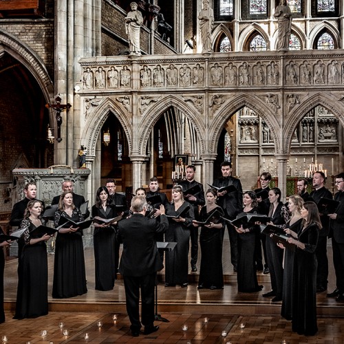 A choir of twenty-one members stand holding binders of music, singing while conducted in an ornate church sanctuary-like room. 