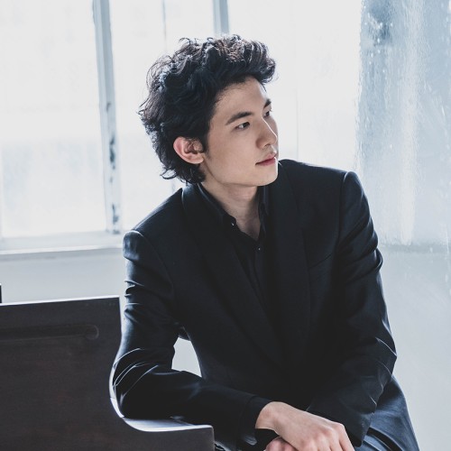 In front of a glassy window, a man with dark, curly hair sits in a black jacket leaning on the lid of the keys on a grand piano. 
