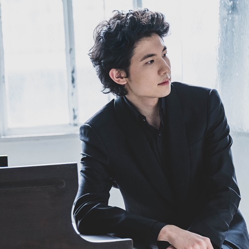 a man in his early 20s, slim and wavy-haired, dressed in a black shirt and jacket jacket, leans on his elbow on a piano