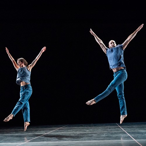 two dancers in blue-green outfits leap in unison -- their straight arms are extended at an angle between their heads and shoulders, and their legs scissor apart in a perpendicular plane from their arms