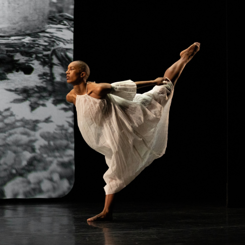 A dancer in sheer white dresses step forward on one foot in front of a black and white photography backdrop