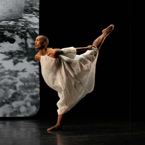 A dancer in sheer white dresses step forward on one foot in front of a black and white photography backdrop