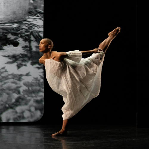 A dancer in sheer white dresses step forward on one foot in front of a black and white photography backdrop