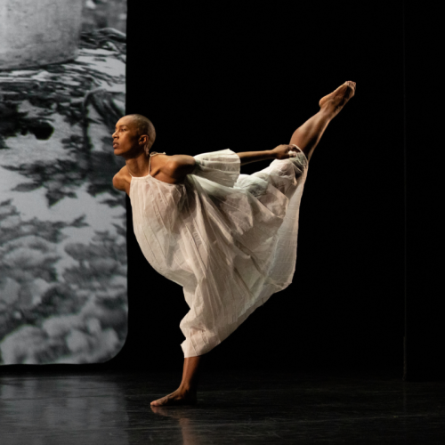 A dancer in sheer white dresses step forward on one foot in front of a black and white photography backdrop
