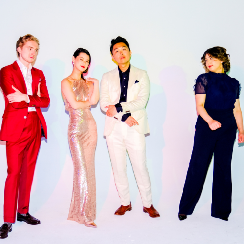 The members of a string quartet wear bold, elegant outfits, and stand before a bright backdrop under colorful light that casts multiple shadows.