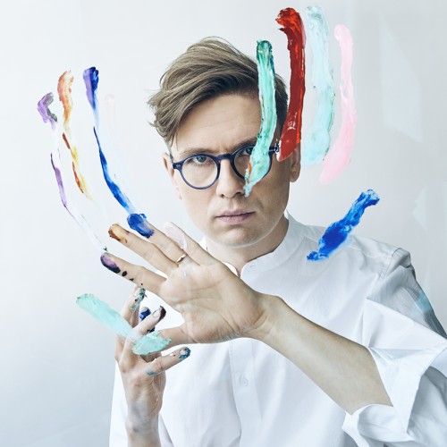 A white man with small round glasses stares intensely ahead while using finger paint on a plexiglass panel in front of his face. 