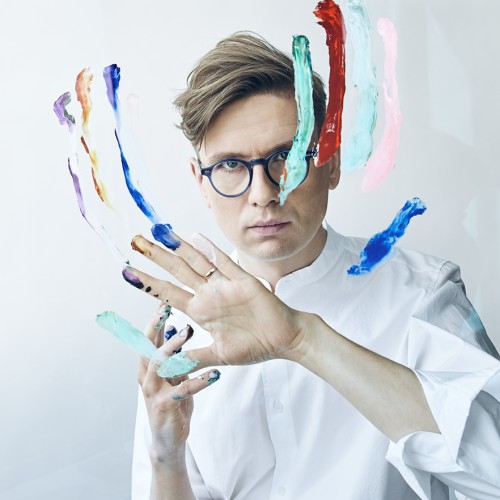 Vikingur Olafsson, a white man with small round glasses, stares intensely ahead while using finger paint on a plexiglass panel in front of his face. 