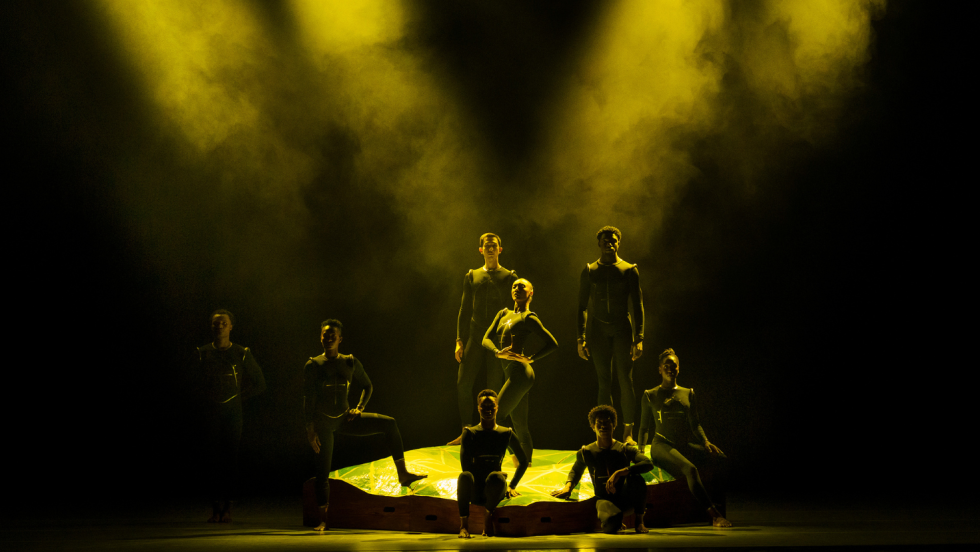 yellow beams of light, defined by haze, cross the stage, barely touching a group of dancers arrayed in a triangle.