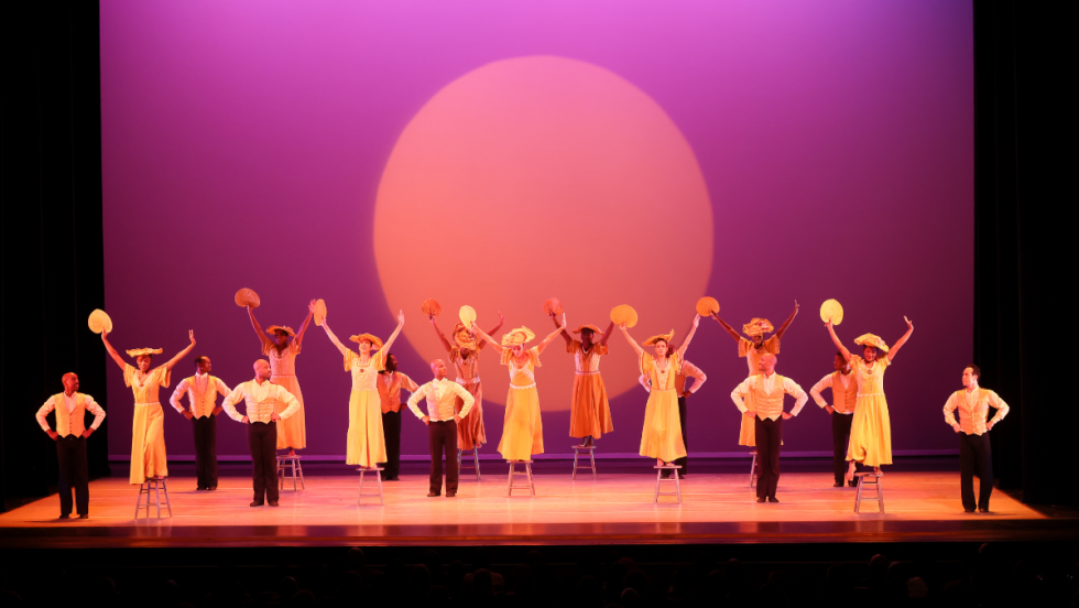 a diverse company of dancers, women in yellow dresses and broad yellow hats who are jumping down from chairs and men in golden vests and dark pants beside them. a setting sun is projected behind them in a warm purple sky.