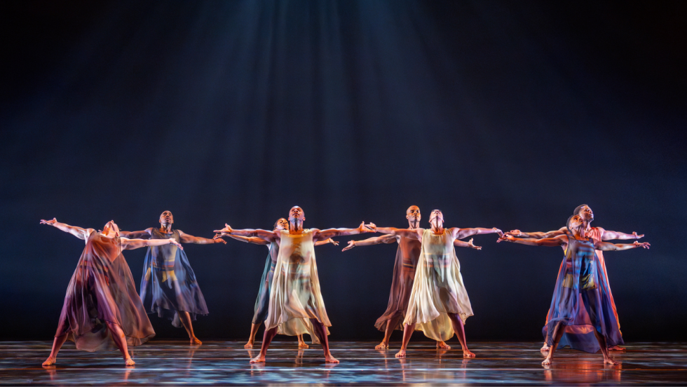 eight dancers in two rows, men and women in flowing sheer gowns, span the stage, arms outstretched. Above them, blue light, defined by haze, radiates in beams from a central point. 