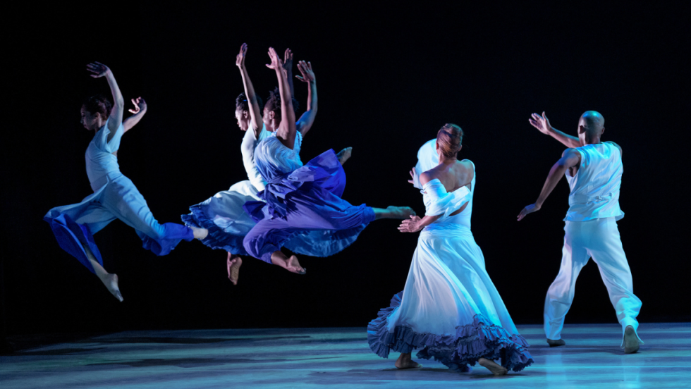 women in blue and white dresses arc their arms up and leap away from us. two dancers in white stride away, crossing their arms across their bodies