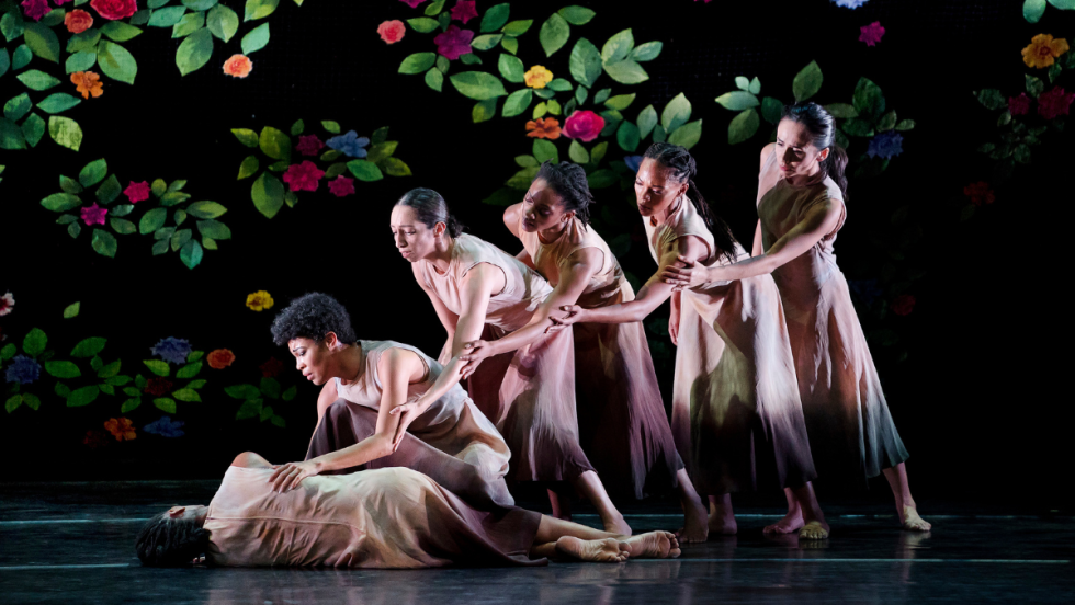 against a black backdrop with vivid painted flowers, a woman lies on her side. Other women lean towards her in a line, each one cupping the elbow of the dancer in front of her.