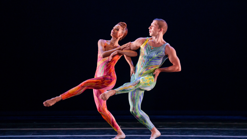a woman in a sparkly pink bodysuit and a man in a sparkly blue-green bodysuit prance arm in arm