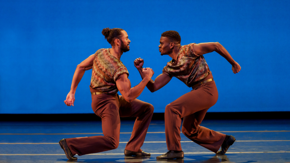 two dancers, both black men, mirror each other's posture of what looks like solidarity. they are in a powerful bent-leg crouch, each with a fist raised on a right-angled elbow toward the other.