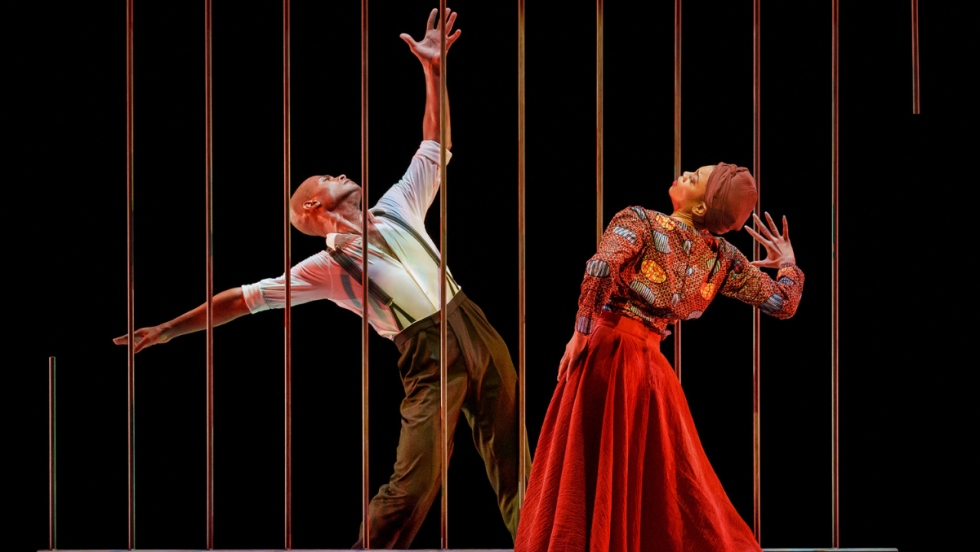a male dancer, depicting nelson mandela, behind jail bars. he reaches and looks upward. a female dancer depicting winnie mandela bends backward, a hand to her forehead 