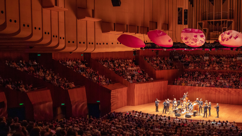 a high-ceiliinged, contemporary, curving, wood-paneled concert hall with round red hanging fixtures above the stage. a chamber orchestra with two guest artists performs.