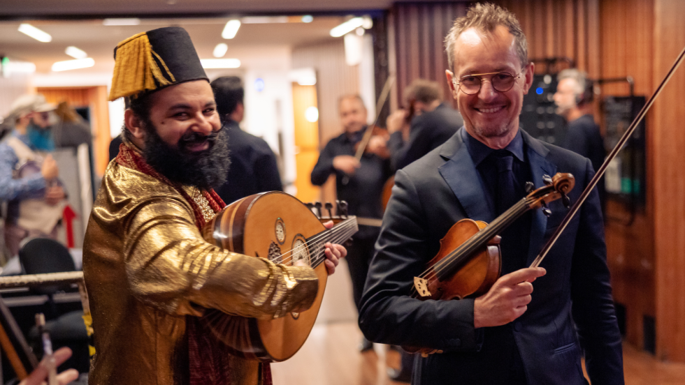 a candid photo of two men smiling. An egyptian australian man in a vivid golden yellow top and a black tasseled hat, turned toward a white man in concert black attire holding a violin under his arm. 