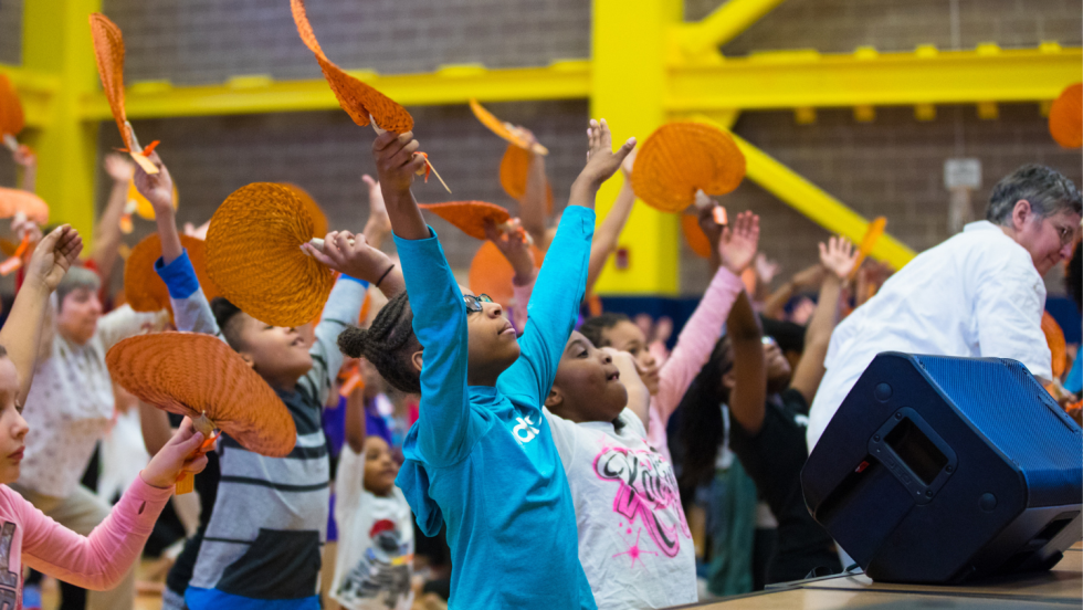 in the first row of an audience before an elevated stage, young black girls raise their arms and join the dance class.