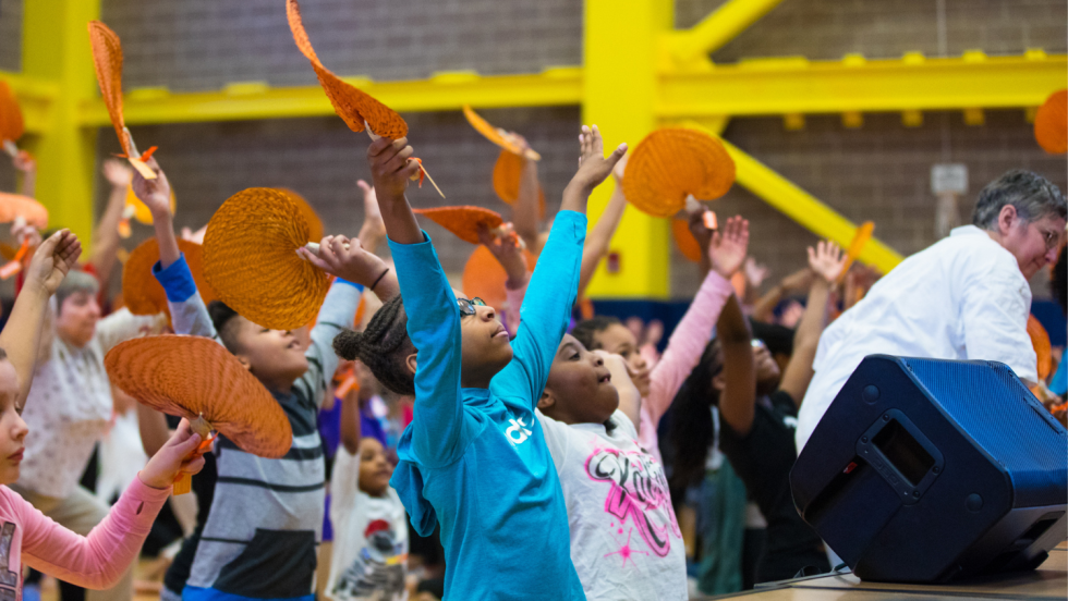 in the first row of an audience before an elevated stage, young black girls raise their arms and join the dance class.