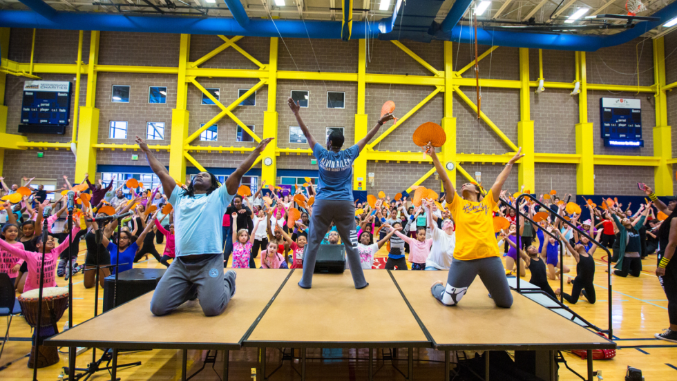 dance instructors on a platform, the central figure stands facing the audience, the others face us, kneeling with arms uplifted.