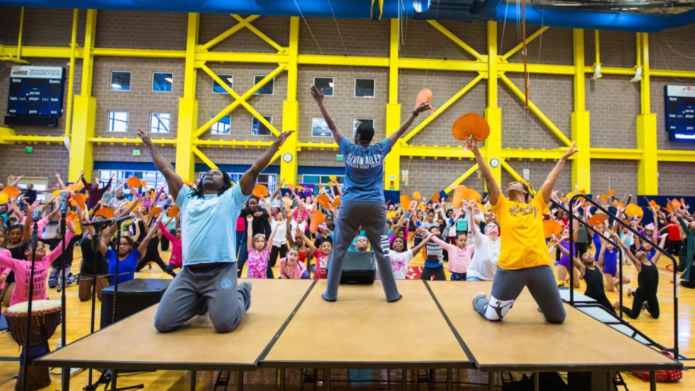 dance instructors on a platform, the central figure stands facing the audience, the others face us, kneeling with arms uplifted.