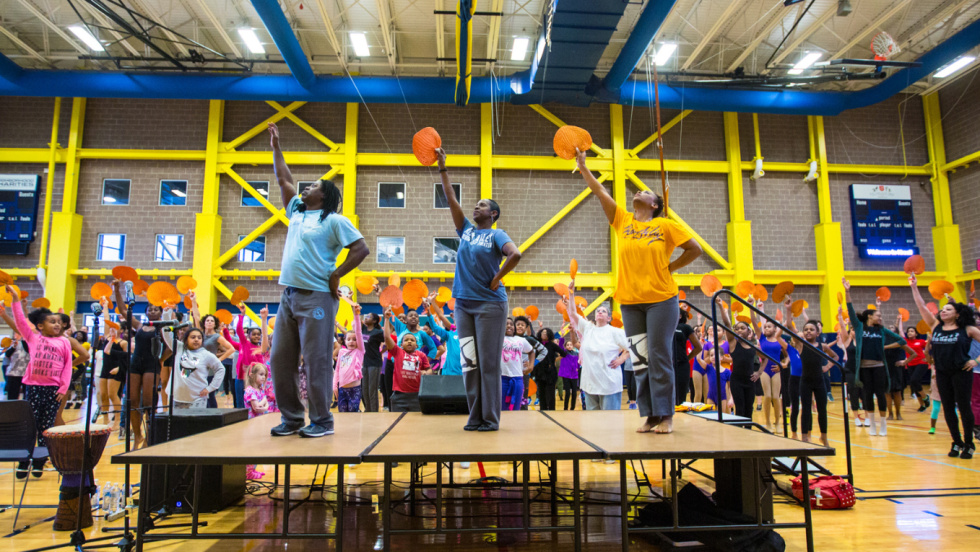 three dance teachers on a platform face away from their audience. all raise fans, all pointing to up and to their right