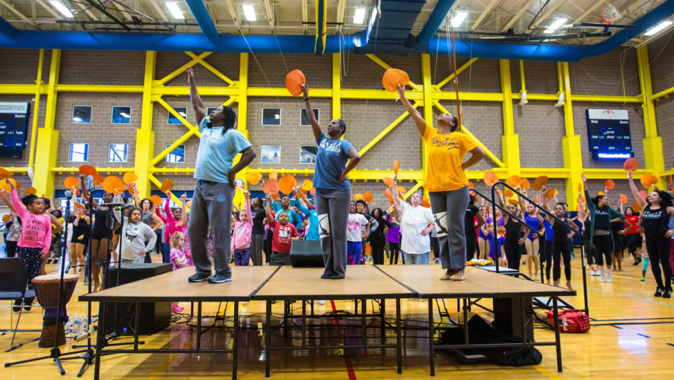 three dance teachers on a platform face away from their audience. all raise fans, all pointing to up and to their right